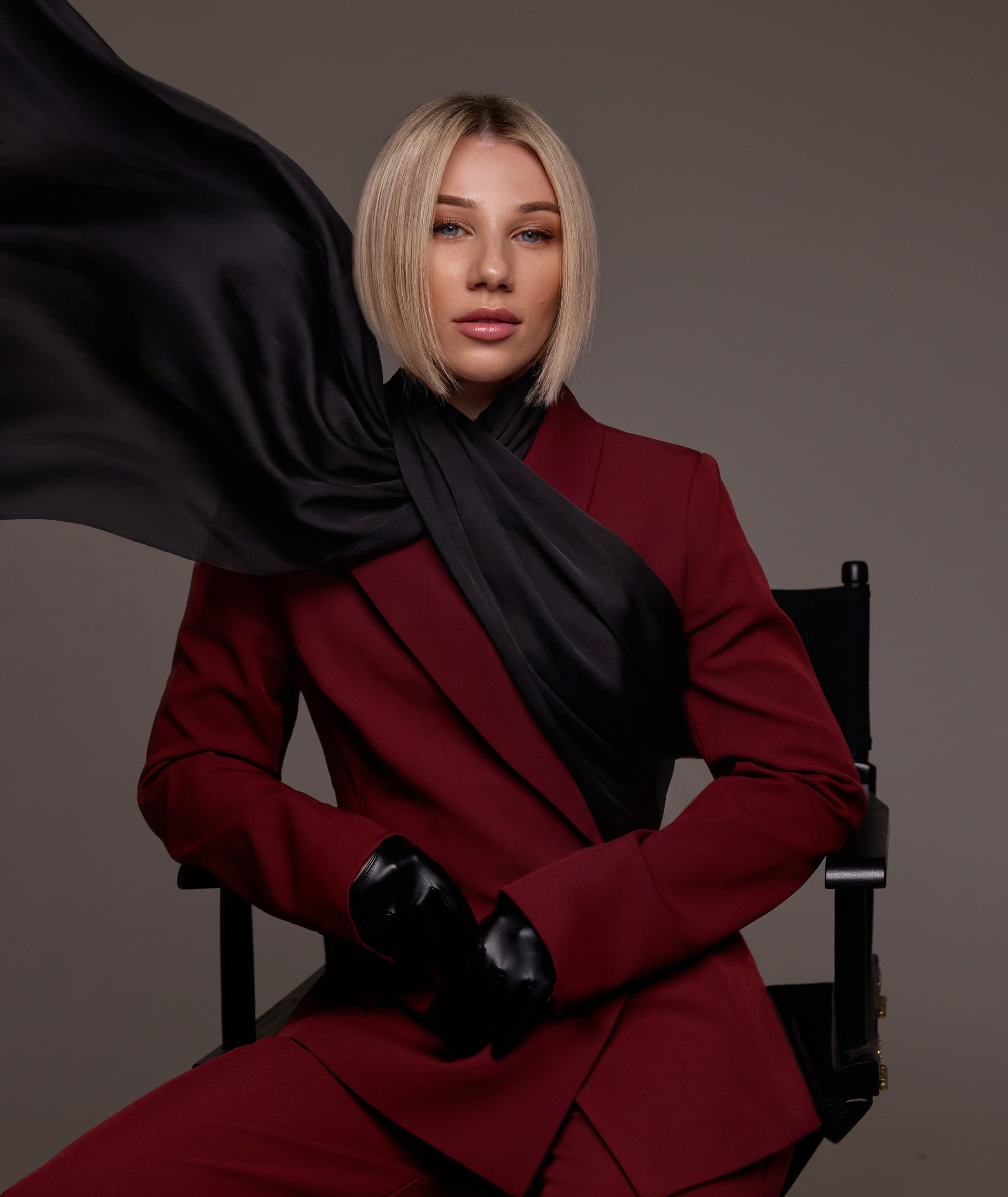 Irma Lion seated in a director's chair wearing a burgundy suit, a black silk scarf in motion